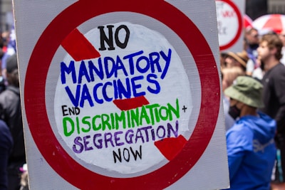 A protest sign with bold, colorful text conveying opposition to mandatory vaccines and a call to end discrimination and segregation. The sign is framed by a red circle with diagonal slashes. In the background, a crowd of people can be seen, some of whom are wearing masks.