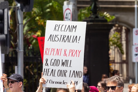 A group of people are gathered outdoors, holding up a sign with the text 'RECLAIM AUSTRALIA! REPENT & PRAY & GOD WILL HEAL OUR LAND 2 Chronicles 7:14'. Some participants are wearing sunglasses and caps. In the background, there is foliage and a traffic light, indicating a city environment.