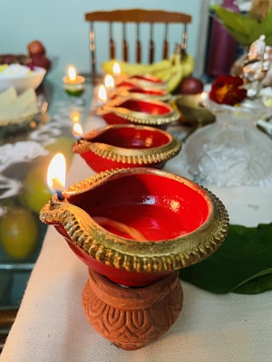 A row of traditional clay lamps with golden rims are lit on a decorated surface, likely for a festive or religious occasion. In the background, there are fruits, a green leaf, and an idol, adding a cultural touch to the setting.