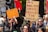 A group of people are participating in a protest, holding up signs with messages. One sign reads 'Discrimination is never ok,' another says '+Safety + Extremism is a threat!,' and a third sign reads 'Jail Dan for crimes against the people.' The protesters are gathered on a street with traffic lights and buildings in the background. The atmosphere appears lively and the individuals express various concerns through their signs.