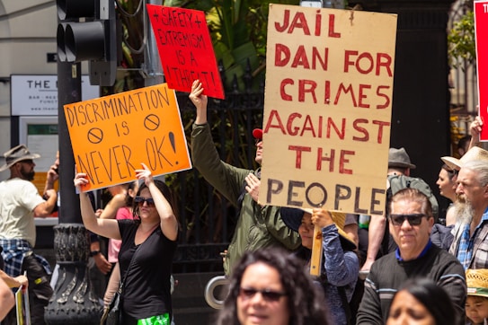 A group of people are participating in a protest, holding up signs with messages. One sign reads 'Discrimination is never ok,' another says '+Safety + Extremism is a threat!,' and a third sign reads 'Jail Dan for crimes against the people.' The protesters are gathered on a street with traffic lights and buildings in the background. The atmosphere appears lively and the individuals express various concerns through their signs.