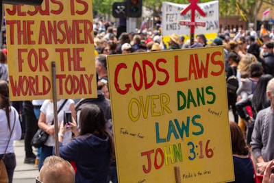 A large group of people gathered at a protest or rally, holding signs with messages related to religious and political beliefs. The signs contain bold letters with slogans such as 'JESUS IS THE ANSWER FOR THE WORLD TODAY' and 'GODS LAWS OVER DANS LAWS.' The crowd appears to be closely packed, with individuals of various ages and appearances.