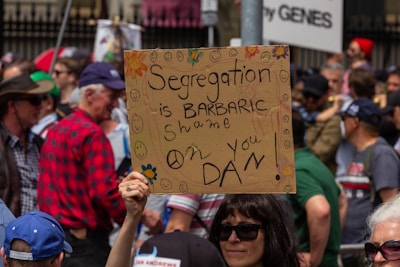 A protest scene features a diverse crowd holding signs. In the foreground, a sign reads 'Segregation is Barbaric shame on you Dan' with smiley faces and peace symbols drawn around it. The participants appear engaged and focused.
