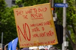 A protest sign made from cardboard is held up, displaying bold red text opposing a surveillance system, digital ID, and social credit system. It is surrounded by trees, people, and a street sign.