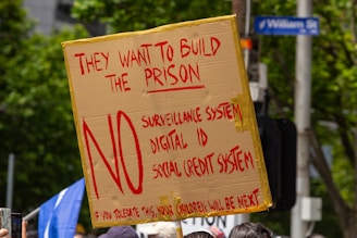 A protest sign made from cardboard is held up, displaying bold red text opposing a surveillance system, digital ID, and social credit system. It is surrounded by trees, people, and a street sign.