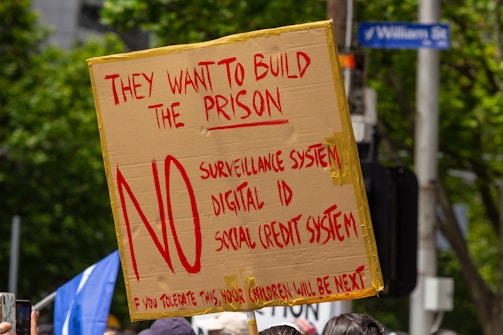 A protest sign made from cardboard is held up, displaying bold red text opposing a surveillance system, digital ID, and social credit system. It is surrounded by trees, people, and a street sign.