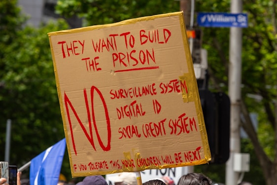 A protest sign made from cardboard is held up, displaying bold red text opposing a surveillance system, digital ID, and social credit system. It is surrounded by trees, people, and a street sign.