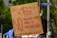 A protest sign made from cardboard is held up, displaying bold red text opposing a surveillance system, digital ID, and social credit system. It is surrounded by trees, people, and a street sign.