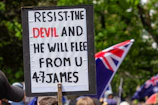 A protest sign with a biblical quote is held up in a crowd. The text on the sign reads, 'RESIST THE DEVIL AND HE WILL FLEE FROM U, 4:7 JAMES.' In the background, several flags with red, white, and blue colors are visible, along with a gathering of people. The scene is set outdoors with greenery surrounding the area.