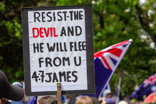 A protest sign with a biblical quote is held up in a crowd. The text on the sign reads, 'RESIST THE DEVIL AND HE WILL FLEE FROM U, 4:7 JAMES.' In the background, several flags with red, white, and blue colors are visible, along with a gathering of people. The scene is set outdoors with greenery surrounding the area.