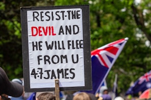 A protest sign with a biblical quote is held up in a crowd. The text on the sign reads, 'RESIST THE DEVIL AND HE WILL FLEE FROM U, 4:7 JAMES.' In the background, several flags with red, white, and blue colors are visible, along with a gathering of people. The scene is set outdoors with greenery surrounding the area.
