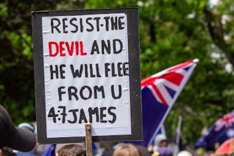 A protest sign with a biblical quote is held up in a crowd. The text on the sign reads, 'RESIST THE DEVIL AND HE WILL FLEE FROM U, 4:7 JAMES.' In the background, several flags with red, white, and blue colors are visible, along with a gathering of people. The scene is set outdoors with greenery surrounding the area.