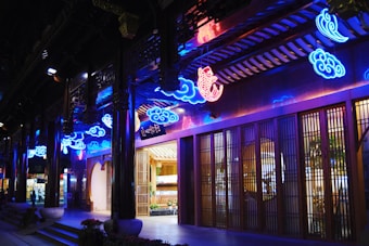 Vibrant neon lights in blue and red hues decorate an Asian-style building facade. The design includes cloud shapes and a prominent fish motif. The structure features wooden doors and traditional architectural elements, illuminated warmly from within.