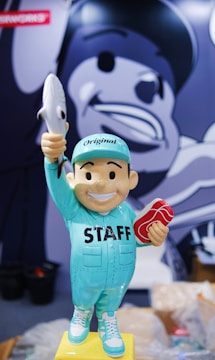A cartoonish statue of a smiling figure wearing a mint green outfit with the word 'STAFF' on the chest, holding a toy shark in one hand and a piece of meat in the other. The figure stands on a yellow base, while a large monochrome mural of a similar face is visible in the background.