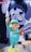 A cartoonish statue of a smiling figure wearing a mint green outfit with the word 'STAFF' on the chest, holding a toy shark in one hand and a piece of meat in the other. The figure stands on a yellow base, while a large monochrome mural of a similar face is visible in the background.
