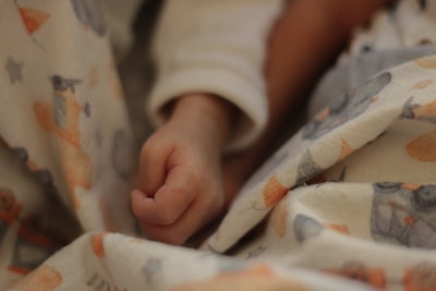 A small infant's hand is partially clenched, resting on soft fabric with a pattern featuring playful animals in muted colors. The setting suggests warmth and tenderness, often associated with a crib or nursery environment.