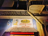 An illuminated sign for a Japanese sushi and sake bar set against a building facade. The sign features a stylized graphic of a woman in traditional Japanese attire, with the establishment's name prominently displayed. Above the awning, there is a decorative architectural element resembling a Greek key pattern. The building is lit warmly, highlighting the colors and textures of the sign and awning.