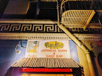An illuminated sign for a Japanese sushi and sake bar set against a building facade. The sign features a stylized graphic of a woman in traditional Japanese attire, with the establishment's name prominently displayed. Above the awning, there is a decorative architectural element resembling a Greek key pattern. The building is lit warmly, highlighting the colors and textures of the sign and awning.