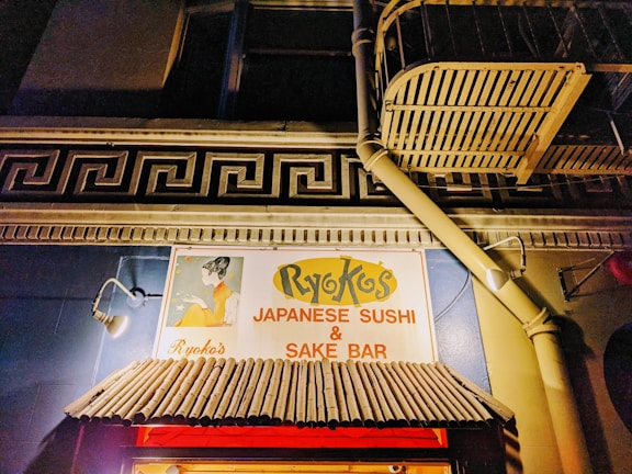 An illuminated sign for a Japanese sushi and sake bar set against a building facade. The sign features a stylized graphic of a woman in traditional Japanese attire, with the establishment's name prominently displayed. Above the awning, there is a decorative architectural element resembling a Greek key pattern. The building is lit warmly, highlighting the colors and textures of the sign and awning.