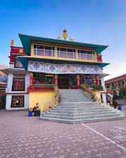 A multi-story traditional building with Tibetan architectural features, including colorful patterns and religious symbols on the facade. The structure has a large staircase leading to the main entrance, adorned with ornamental columns and decorative elements. The roof is elaborately designed with vivid colors and intricate details.