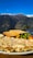 A plate of food including a golden omelette, two slices of toasted bread, and a mix of sautéed vegetables such as bell peppers, broccoli, and carrots. The meal is set against a backdrop of a mountainous landscape with snow-capped peaks and a clear blue sky.