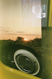 A double exposure image blending a sunset over a field with the close-up of a vintage car's wheel, showcasing intricate textures. The sky is filled with soft clouds, and the warm light of the setting sun creates a serene atmosphere. The car is yellow and parked on dried grass, adding an element of nostalgia and tranquility.