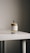 A clear glass bottle of perfume with a spray nozzle is placed on a light-colored surface. The label on the bottle reads 'TONKA 25' and includes other text details.