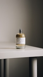 A clear glass bottle of perfume with a spray nozzle is placed on a light-colored surface. The label on the bottle reads 'TONKA 25' and includes other text details.