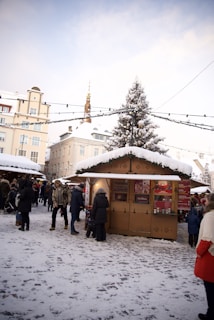 A snowy outdoor market scene with people walking around and visiting stalls. In the foreground, a wooden stall is decorated with festive garlands and snow, offering traditional holiday drinks. Several individuals are dressed in winter clothing, engaged in conversation or purchasing items. A tall Christmas tree is visible in the background, adorned with snow. The sky is clear, and surrounding buildings create a cozy, festive atmosphere.