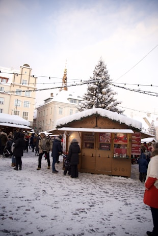 A snowy outdoor market scene with people walking around and visiting stalls. In the foreground, a wooden stall is decorated with festive garlands and snow, offering traditional holiday drinks. Several individuals are dressed in winter clothing, engaged in conversation or purchasing items. A tall Christmas tree is visible in the background, adorned with snow. The sky is clear, and surrounding buildings create a cozy, festive atmosphere.