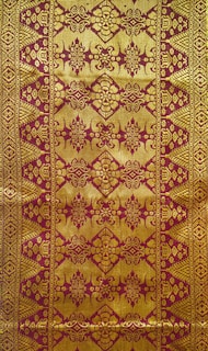 Intricate textile design featuring ornate golden patterns on a reddish-brown background, with symmetrical, repetitive motifs that include floral and geometric elements.