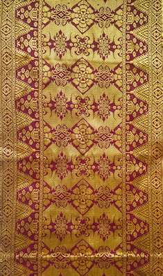 Intricate textile design featuring ornate golden patterns on a reddish-brown background, with symmetrical, repetitive motifs that include floral and geometric elements.