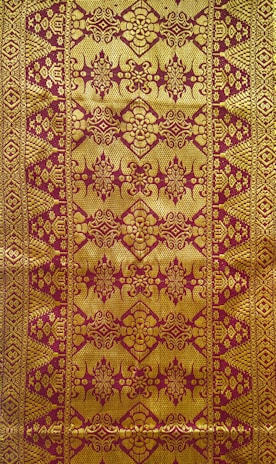 Intricate textile design featuring ornate golden patterns on a reddish-brown background, with symmetrical, repetitive motifs that include floral and geometric elements.