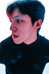 A close-up portrait captures a person with black hair, wearing a black top, against a light background. The lighting casts a slight red tint on the left side of the face, creating a dramatic and artistic effect.