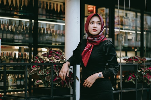 A woman wearing a red hijab and a black outfit is standing near a railing with plants. Behind her, there is a display of wine bottles on shelves, indicating the location might be a wine shop or a bar.