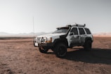 A rugged, mud-covered SUV is parked on a dry, expansive terrain with mountains visible in the distance under a clear sky. The vehicle appears to be equipped for off-road adventures, with a roof rack and heavy-duty tires.