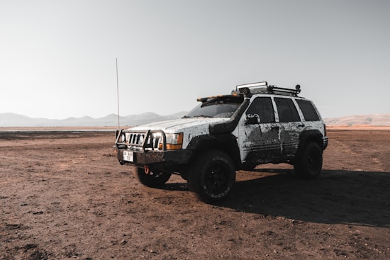 A rugged, mud-covered SUV is parked on a dry, expansive terrain with mountains visible in the distance under a clear sky. The vehicle appears to be equipped for off-road adventures, with a roof rack and heavy-duty tires.