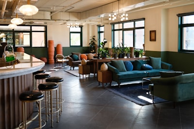 A well-lit, stylish lounge area with large windows and elegant lighting fixtures. The room features plush green sofas arranged around a coffee table, with decorative pillows adding a splash of color. Potted plants line a wooden console table beneath the windows, bringing a touch of nature indoors. The space includes a bar area with black stools and a marble countertop. The floor is tiled in a dark gray, complementing the room's modern decor.