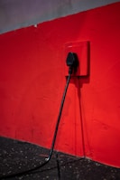 A black electrical plug is inserted into a red wall socket. The wall is painted in a bold red color, and the cord hangs down towards a dark, textured floor.