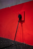 A black electrical plug is inserted into a red wall socket. The wall is painted in a bold red color, and the cord hangs down towards a dark, textured floor.