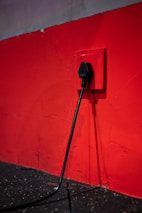 A black electrical plug is inserted into a red wall socket. The wall is painted in a bold red color, and the cord hangs down towards a dark, textured floor.