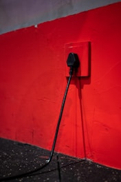 A black electrical plug is inserted into a red wall socket. The wall is painted in a bold red color, and the cord hangs down towards a dark, textured floor.