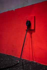 A black electrical plug is inserted into a red wall socket. The wall is painted in a bold red color, and the cord hangs down towards a dark, textured floor.