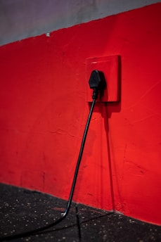 A black electrical plug is inserted into a red wall socket. The wall is painted in a bold red color, and the cord hangs down towards a dark, textured floor.
