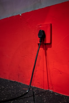 A black electrical plug is inserted into a red wall socket. The wall is painted in a bold red color, and the cord hangs down towards a dark, textured floor.