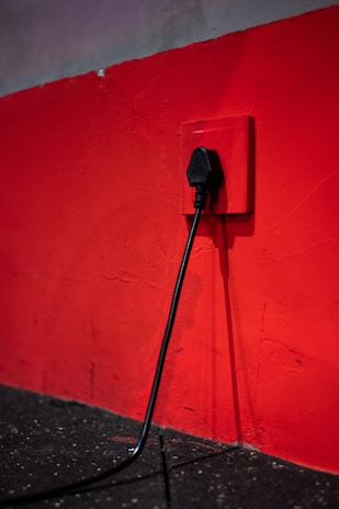 A black electrical plug is inserted into a red wall socket. The wall is painted in a bold red color, and the cord hangs down towards a dark, textured floor.