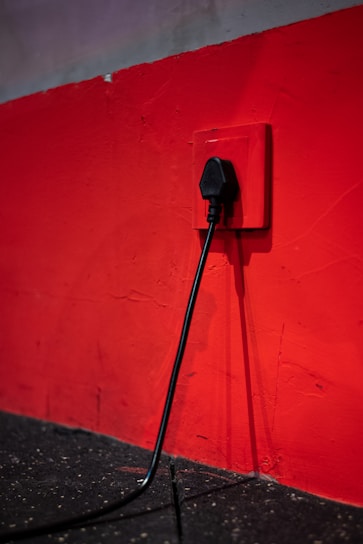A black electrical plug is inserted into a red wall socket. The wall is painted in a bold red color, and the cord hangs down towards a dark, textured floor.
