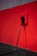 A black electrical plug is inserted into a red wall socket. The wall is painted in a bold red color, and the cord hangs down towards a dark, textured floor.