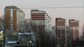 Several high-rise residential buildings are visible, featuring a combination of red brick and white panels. Leafless trees are in the foreground, and there are visible signs with neon lights on the lower level, indicating a commercial area.