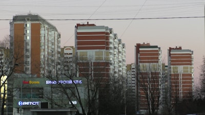 Several high-rise residential buildings are visible, featuring a combination of red brick and white panels. Leafless trees are in the foreground, and there are visible signs with neon lights on the lower level, indicating a commercial area.